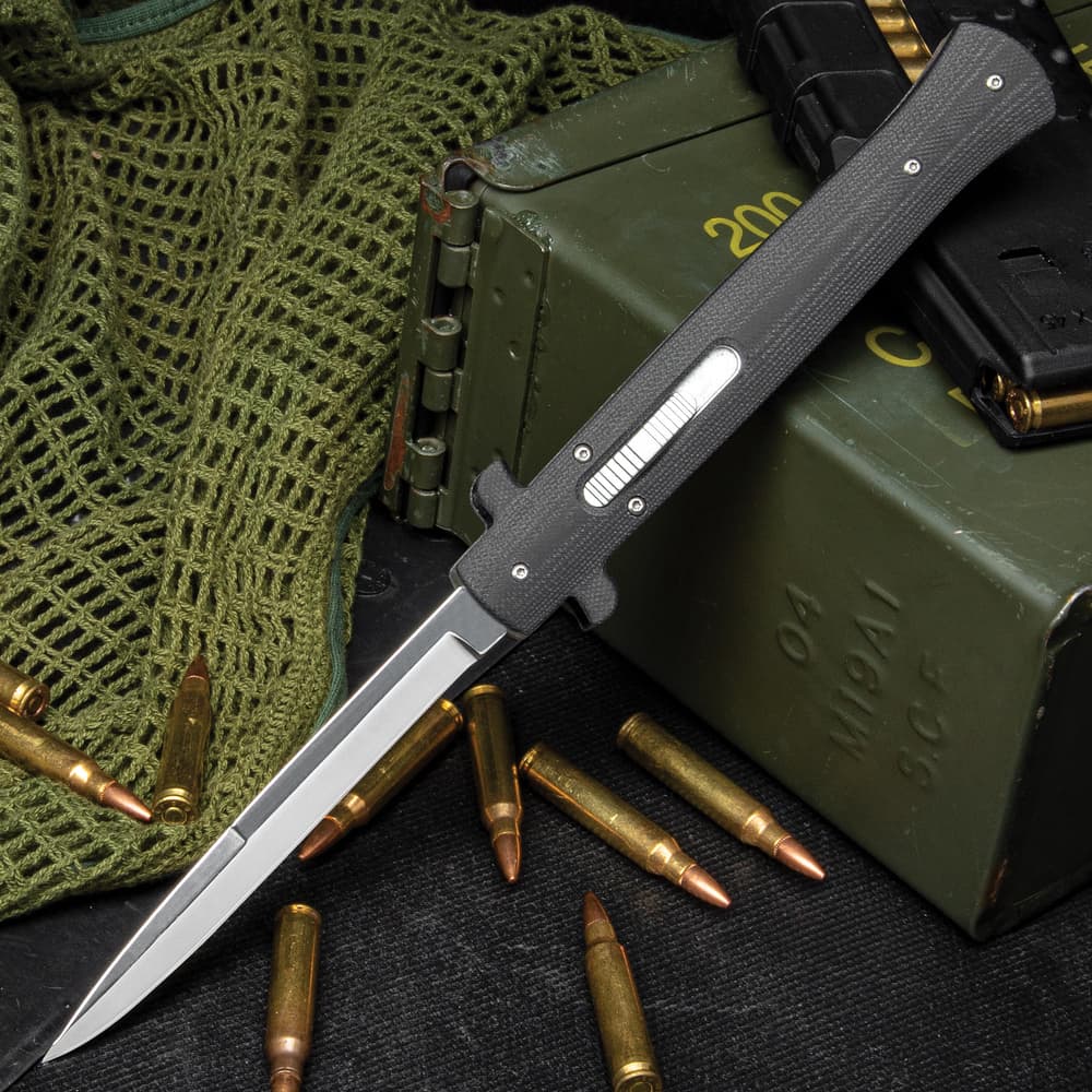 Large OTF automatic stiletto knife with slide button trigger and mirror-polished blade on a background of black and green utility gear. image number 0