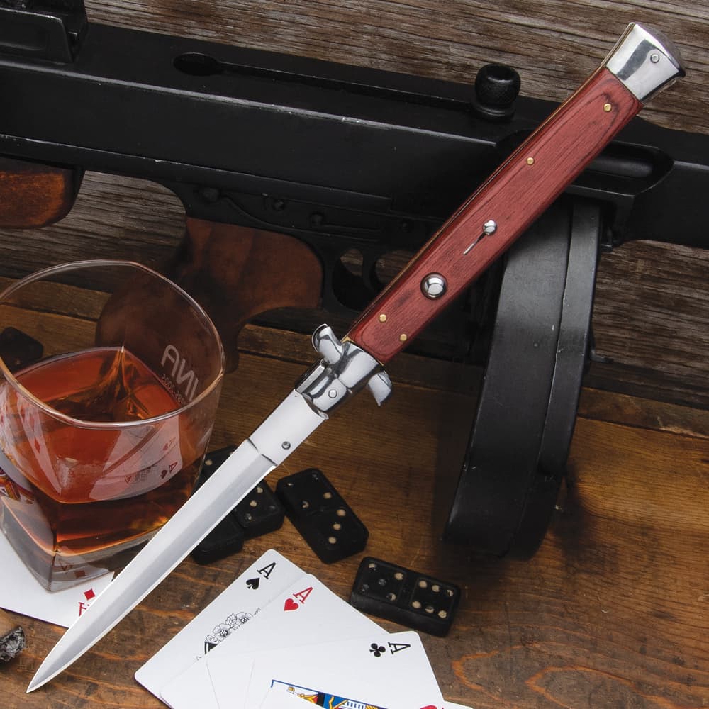 Italian stiletto knife with wooden handle and mirror-polished accents on a background of wood with whiskey, cards, and dominos. image number 0