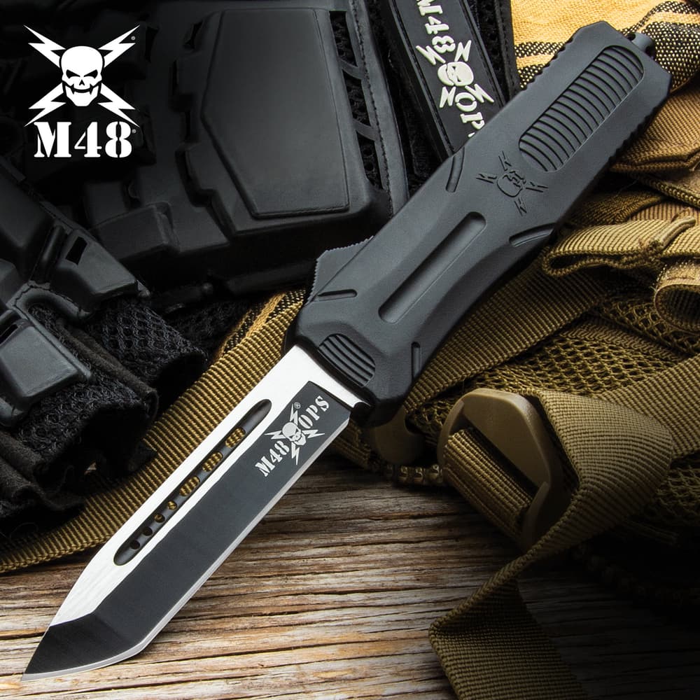 Matte black OTF tanto knife with a glass breaking pommel featuring a "M48" logo on both the blade and the handle on a background of tactical gear. image number 0