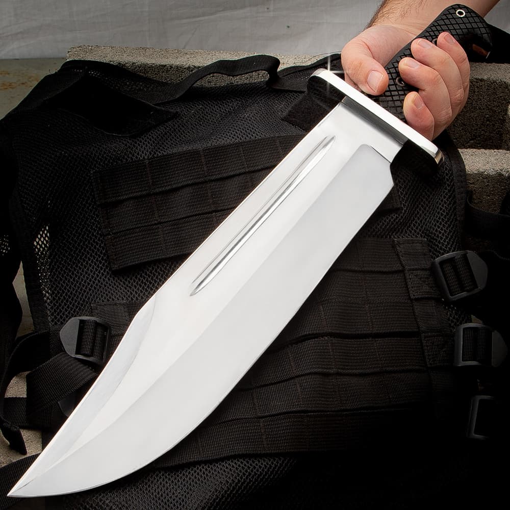 Large shining silver bowie knife being held at a downwards left angle on a background of grey concrete and a black tacticle bag. image number 0