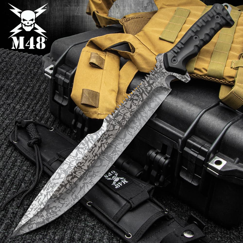 Combat machete with spiderweb titanium electroplated finish and a partial sawback spine on a background of black and tan utility gear. image number 0