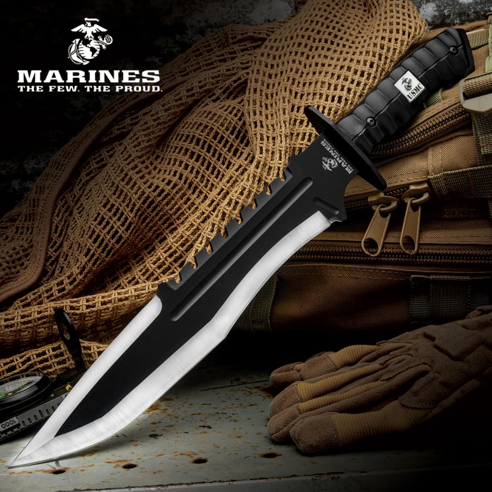 The USMC Bulldog Bowie Knife with two-toned stainless steel blade and black textured handle shown beneath the Marines logo, laid on a tactical backpack. image number 0