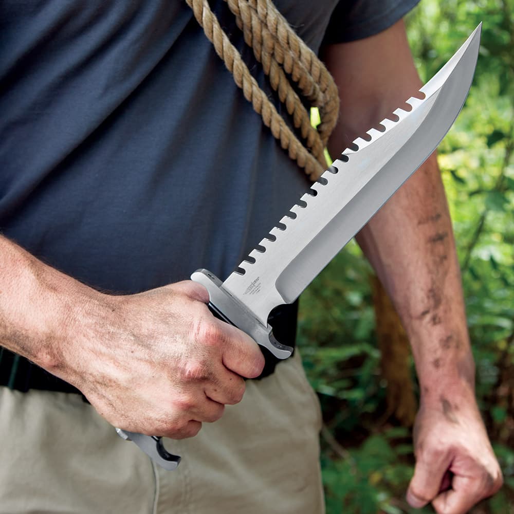 A hand is shown holding the knife by its black pakkawood handle with the sawback stainless steel blade facing upward. image number 1
