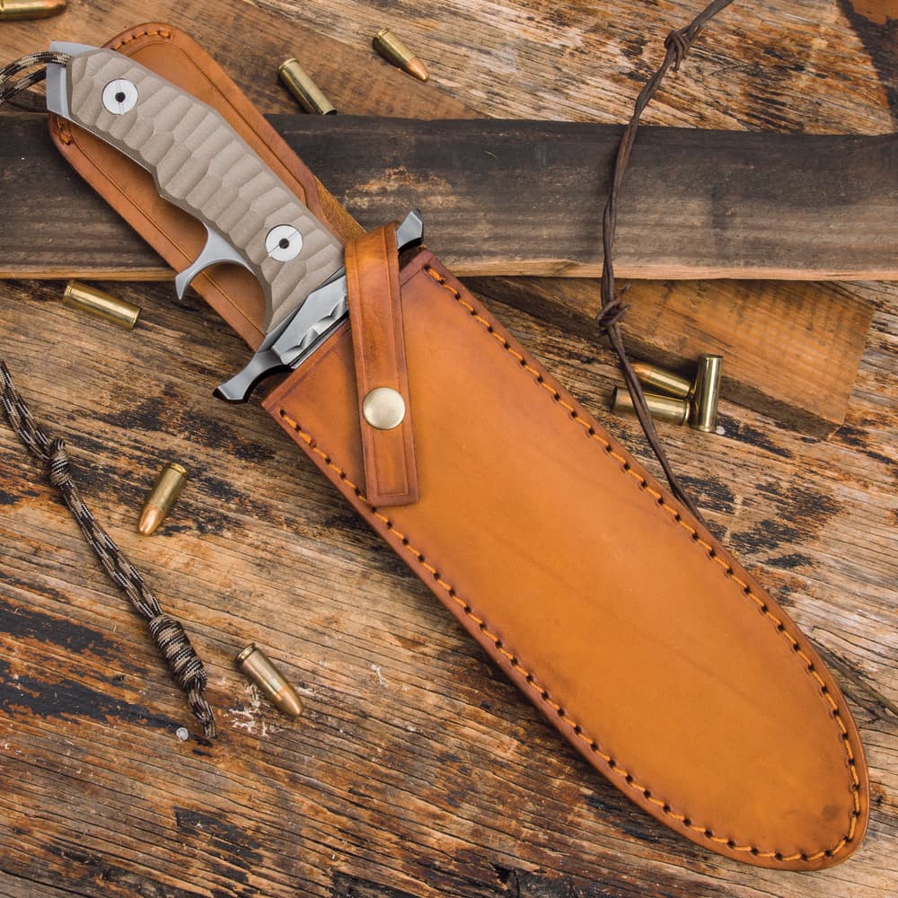 Knife enclosed in chesnut leather sheath with patina and snap closure on a background of burnt wood, barbwire, and shell casings. image number 1