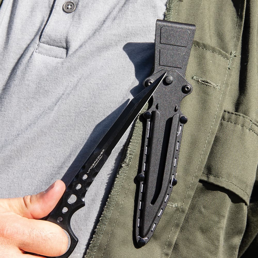 Black "Undercover" stinger knife being held through the open ring pommel in front of black knife sheath attached to utility jacket. image number 1