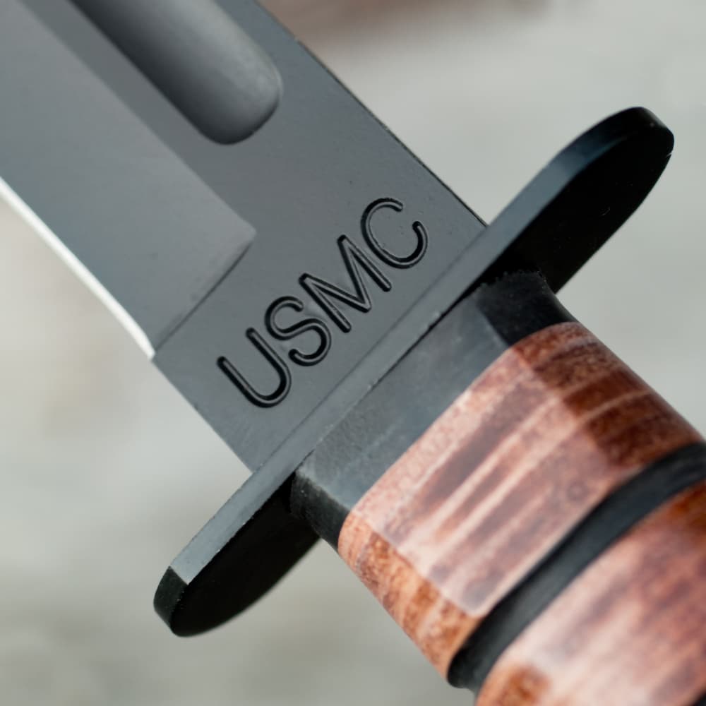 Upclose view of a USMC combat fighter knife with a large engraving of "USMC" above the blood groove and a polished stacked leather handle. image number 2