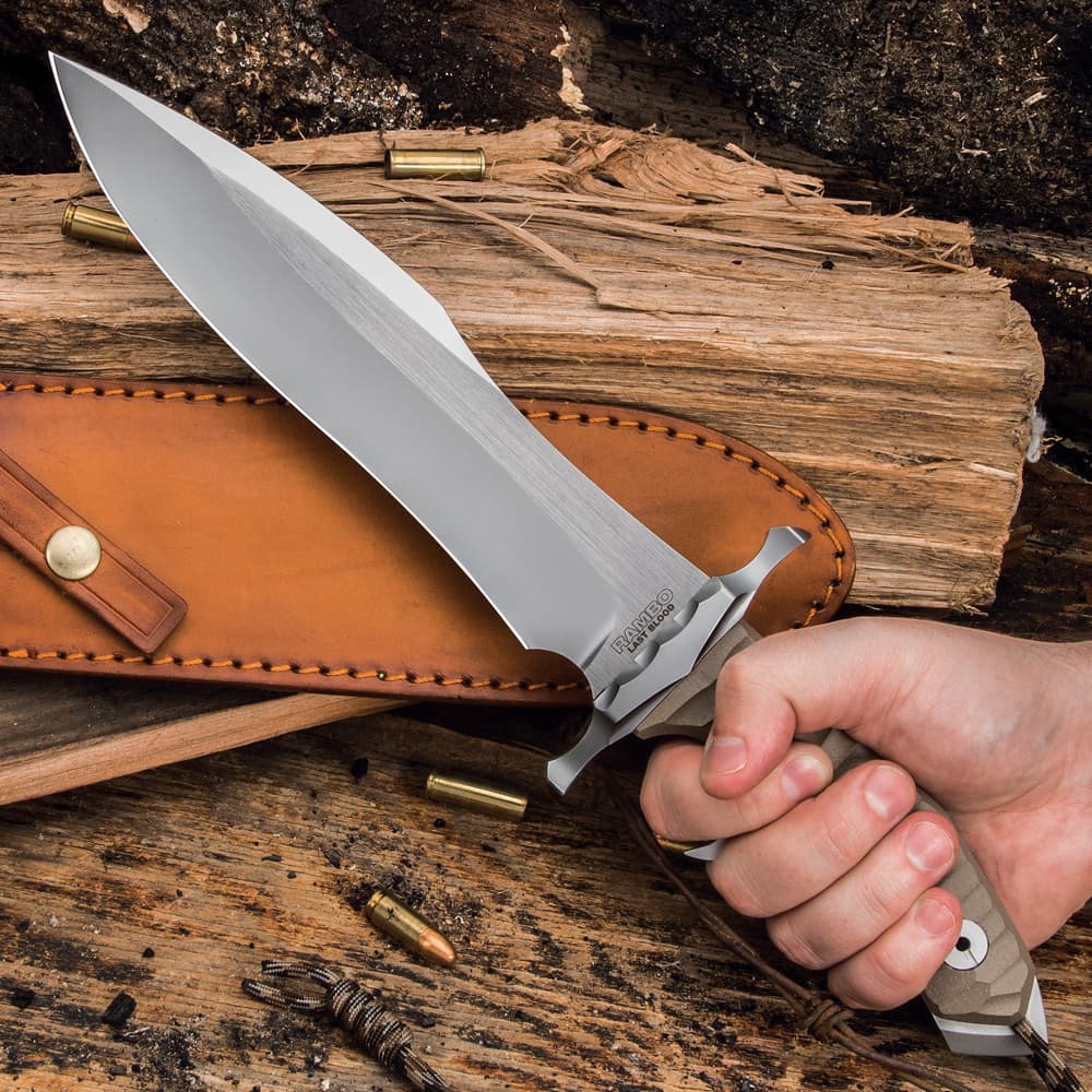 Non-reflective "Rambo" knife being held in front of leather sheath on a back ground of wood and shell casings. image number 2