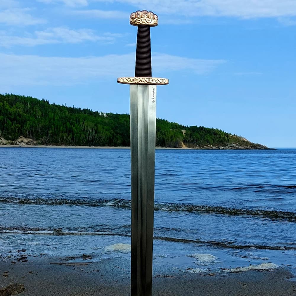 The Ballinderry Viking Sword is shown in front of a body of water, showcasing the unique handle and 1065 carbon steel blade. image number 2