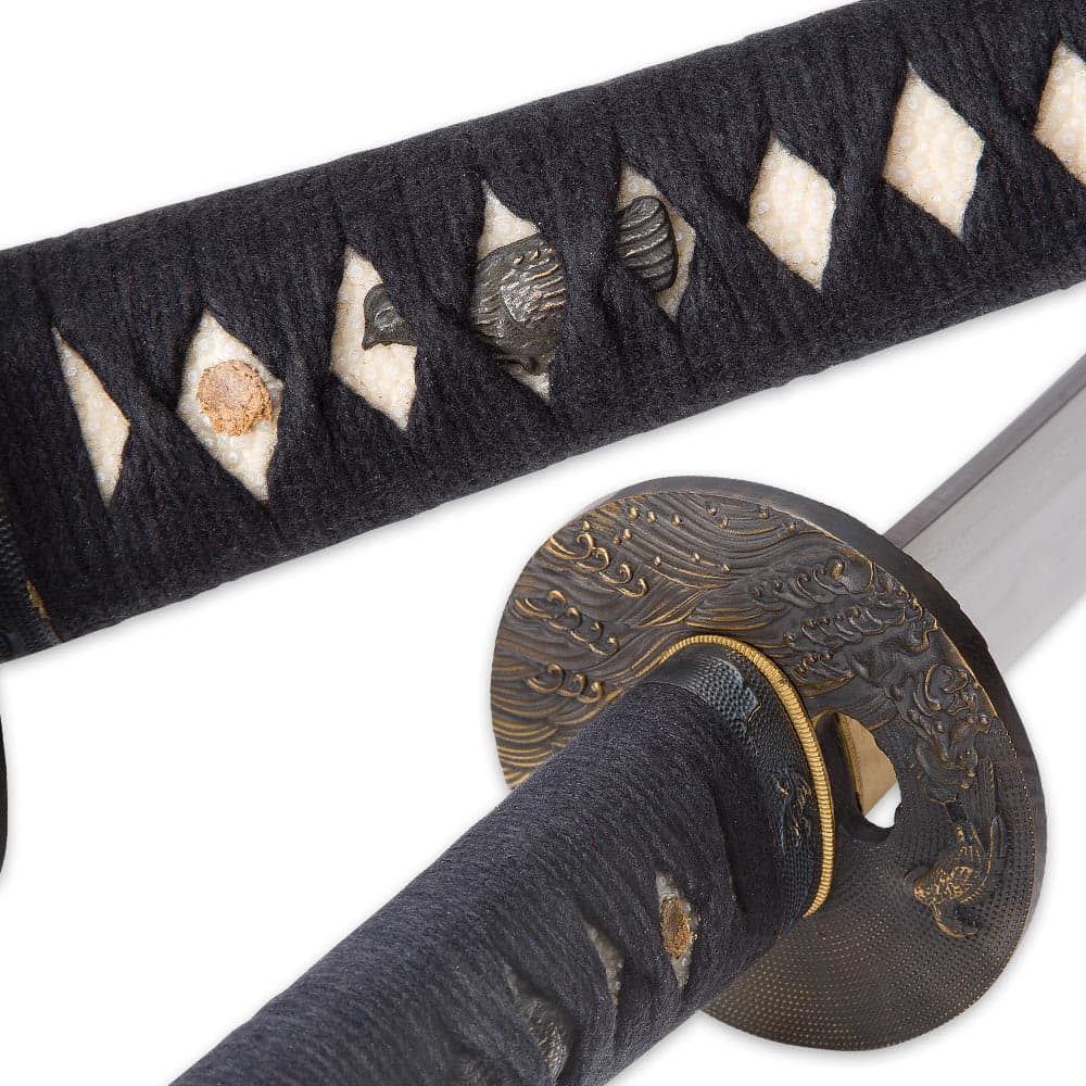 A bird menuki peaks out from behind the black braided cord wrapping on the ray skin handle next to view of the brass tsuba. image number 2