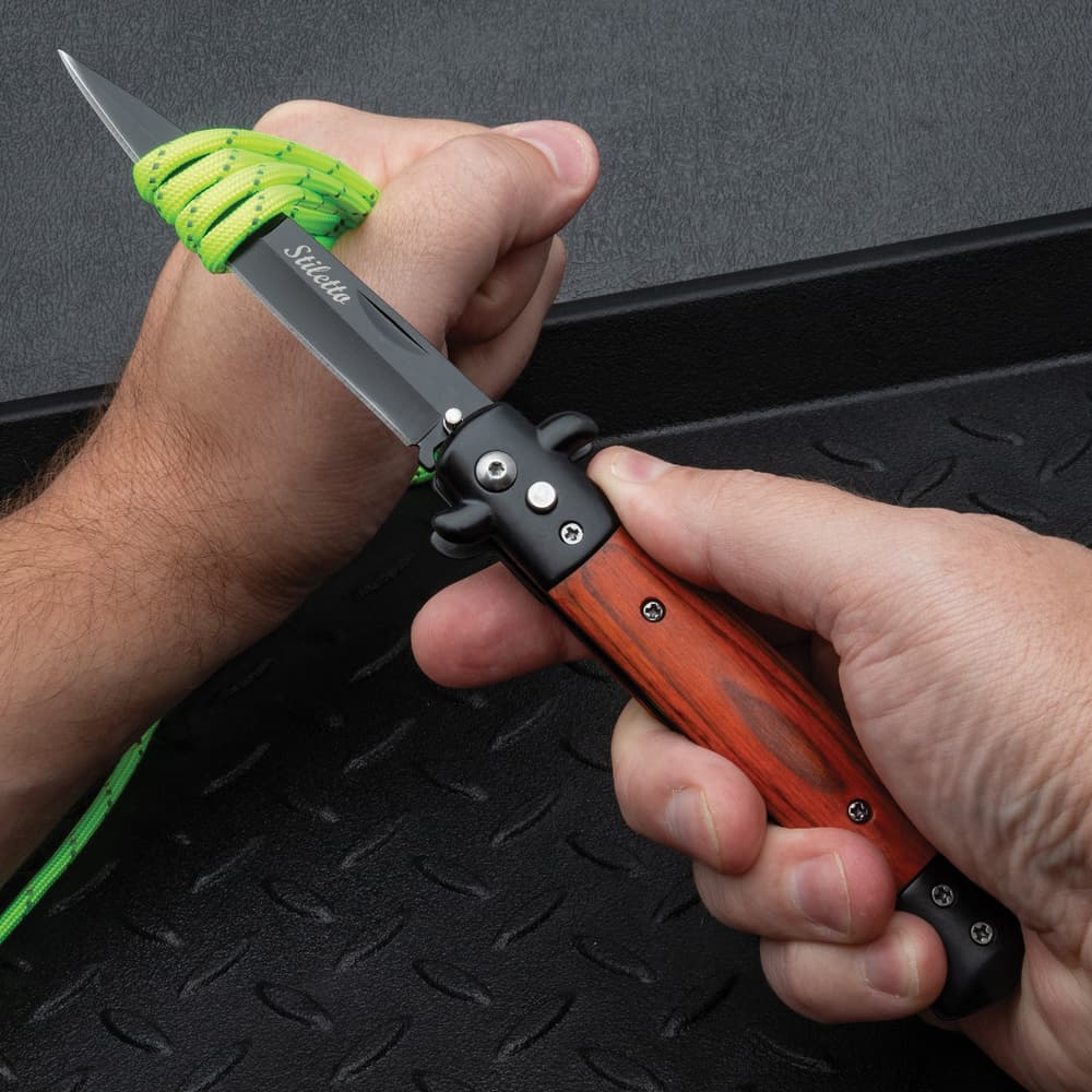 The Gangster's Edge Automatic Stiletto Pocket Knife in action, effortlessly cutting through green cord with its 3 7/8" black coated stainless steel blade. image number 3