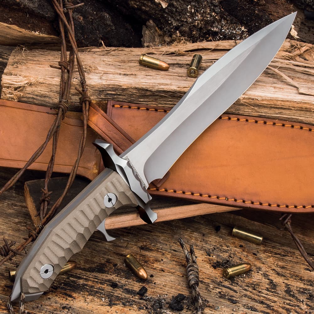 Angled knife with taupe textured handle and stainless steel blade resting on leather sheath and wood background and scattered bullets. image number 4