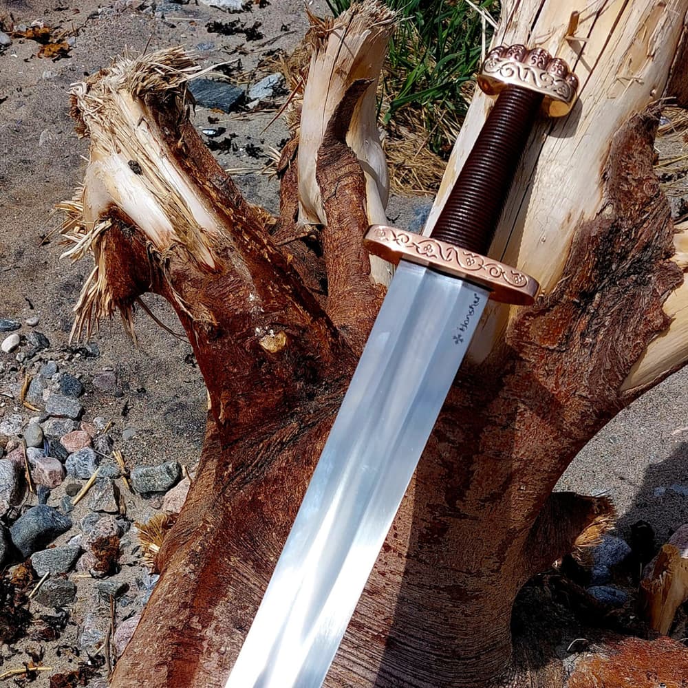 The Honshu Ballinderry Viking Sword shown against a tree with view of the brass pommel and guard and brown leather wrapped handle. image number 4