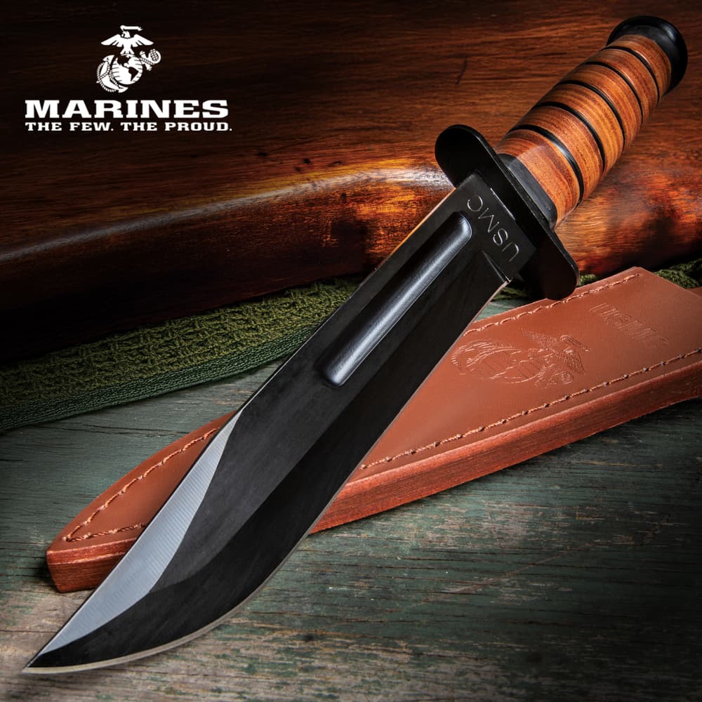 Fixed blade knife with stacked leather handle and large "USMC" engraving on a green antique wood background with a leather sheath and wooden gun handle. image number 0