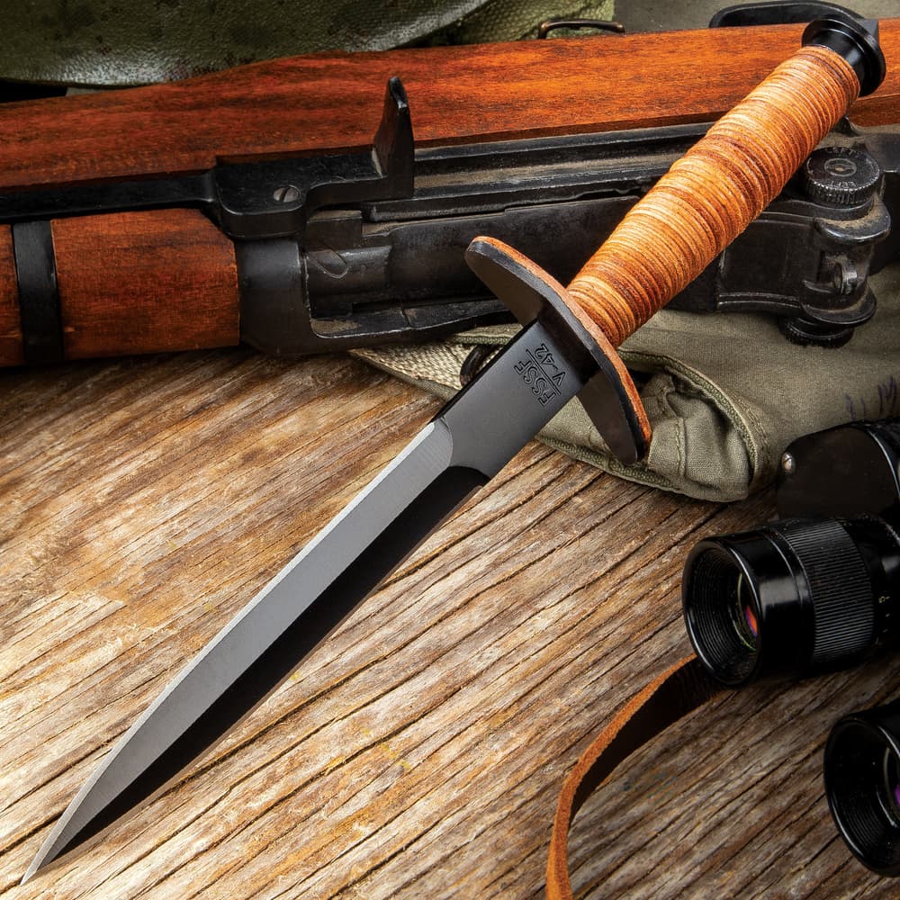 Stiletto dagger with a black blade finish and stacked leather handle on a background of wood and tactical gear. image number 0