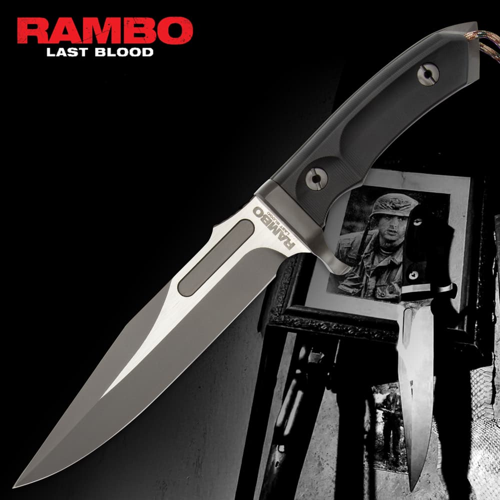 Rambo Last Blood Bowie Knife rests on a black leather sheath. It features a satin-finish blade with a bold “RAMBO” engraving near the hilt. The textured handle is secured by a large polished screw image number 0