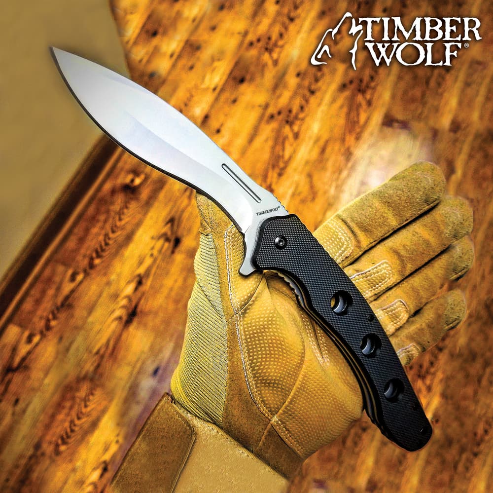 Gloved hand holding a Timber Wolf knife with a black handle on a wooden floor background image number 0