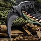 The 7 1/2” karambit has a 3 1/2” sharpened blade with fierce curve shown on a background of tactical gear.