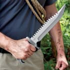 A hand is shown holding the knife by its black pakkawood handle with the sawback stainless steel blade facing upward.