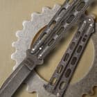 Detailed view of the intricate Damascus steel blade and dark epoxy powder-coated handles on a metal background.