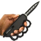 A hand is shown holding the knuckle handle with a finger deploying the blade using the slide on the spine.