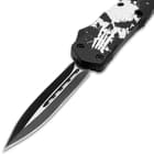 Upclose view of an open OTF pocket knife with a matte black and silver dual toned dagger style blade and a blade handle with a large white distressed skull print.