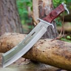 This knife has a 12” stainless steel machete style blade and dark pakkawood handle, shown outside on a piece of wood.