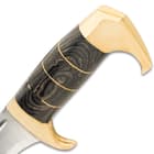 Th handle is crafted of grey pakkawood, accented with brass spacers, and the hooked pommel is of polished brass