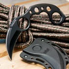 The black karambit with finger holes and ribbed details measures 6 1/2" overall and is shown next to its black plastic sheath on a wooden background.
