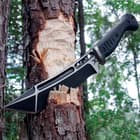 The knife’s black stainless steel blade is shown sliced into the side of a tree.