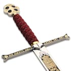 Zoomed view of the black and gold detailing of the blade and guard, beneath the red handle with gold cord wrapping. 