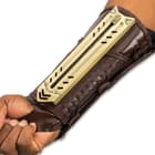Retracted tactical gauntlet compossed of mahogany brown faux leather and gold blade enclosure attached to the inner forearm.
