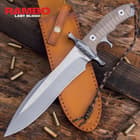 Stainless steel knife with hefty hand guard and tungsten carbide coating resting upon a chesnut leather sheath, with a wood background and scattered bullets.

