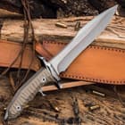 Angled knife with taupe textured handle and stainless steel blade resting on leather sheath and wood background and scattered bullets. 

