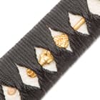Close view of the brass menuki on the white ray skin handle with black braided cord. 