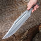 Large "Gil Hibben" mirror polished throwing knife being held diagonally in front of a wood background. 
