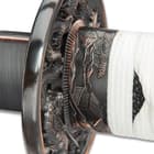 Zoomed in view of habaki and tsuba with embossings extended to faux rayskin handle wrapped with white cord
