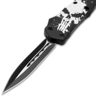 Upclose view of an open OTF pocket knife with a matte black and silver dual toned dagger style blade and a blade handle with a large white distressed skull print.