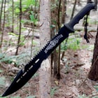The machete’s black coated blade with cutouts and sawback blade design is shown stuck into a tree trunk.