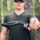 A man is shown holding the kukri by the black TPR handle with the curved black stainless steel blade in full view with the M48 logo just beneath the guard.