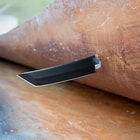 The 1065 carbon steel blade with black hard coating is shown piercing through a piece of material. 