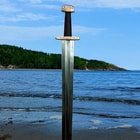 The Ballinderry Viking Sword is shown in front of a body of water, showcasing the unique handle and 1065 carbon steel blade.