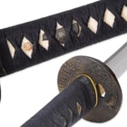 A bird menuki peaks out from behind the black braided cord wrapping on the ray skin handle next to view of the brass tsuba. 