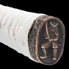 The handle is wrapped in gold-threaded cloth and white cord and has a cast metal pommel with a Samurai theme