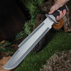 A hand is shown holding the machete by its black pakkawood handle outside with the stainless steel blade in full view.