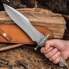 Stainless steel knife with hefty hand guard and tungsten carbide coating resting upon a chesnut leather sheath, with a wood background and scattered bullets.
