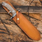 Knife enclosed in chesnut leather sheath with patina and snap closure on a background of burnt wood, barbwire, and shell casings. 
