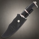 Large knife with black, red, and silver, hilt exposed with the blade enclosed in a black leather sheath stamped with "Hibben Knives" logo 
