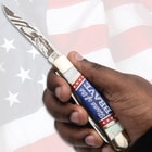 Hand holding knife with 'Home of the Brave' handle on flag