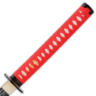 The hardwood handle is wrapped in faux rayskin and red cord and has an intricately detailed metal alloy tsuba
