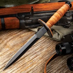 Stiletto dagger with a black blade finish and stacked leather handle on a background of wood and tactical gear.